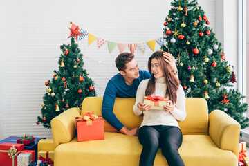 Young Asian man surprises her girlfriend that wearing Santa Claus hat with a Christmas gift at home with a Christmas tree in the background. Image with copy space.