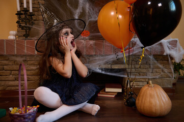 Surprised Caucasian little girl in witch black dress and wizard hat, looks at balloons, sitting near pumpkin and basket with candies against a fireplace covered with spider web. Happy Halloween party