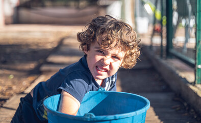 Child makes a grimace looking at the camera while playing, putting his hand inside a bucket of water, in a garden of a rural house.