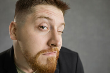 Closeup studio shot of funny curious bearded young man looking at you with one eye with expression of suspicion, skepticism, doubt and disbelief on face isolated on grey background