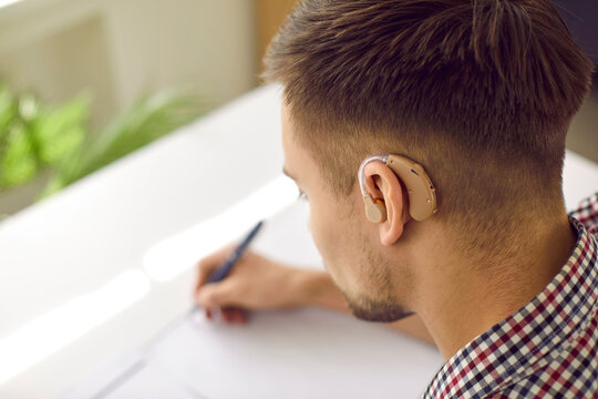 Student with hearing aid doing homework. Young deaf man wearing plastic hearing device behind his ear sitting at desk and writing. Close up of man's head. Hearing loss, impairment, education concept