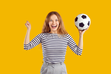 Happy funny pretty woman standing isolated on yellow background, holding soccer ball and cheering. Excited beautiful young girl supporting her favorite soccer team or celebrating their victory