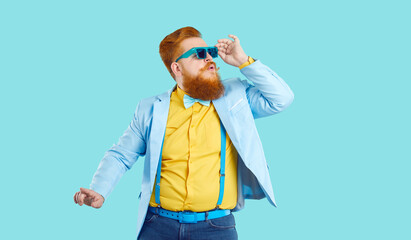 Fat young man in funky modern outfit having fun at disco party. Funny fashion guy with ginger beard and mustache wearing trendy blue specs, jacket, yellow shirt and bowtie dancing on blue background - obrazy, fototapety, plakaty