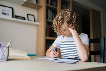 Portrait of pensive little child schoolgirl writing in exercise book doing homework, sitting at desk near window. Cute curly small school girl studying writing task, taking notes, learning at home.