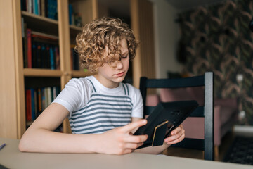Close-up of upset preteen girl holding photo frame in hands and looking sorrowfully at family portrait. Frustrated adorable kid missed person in picture. Concept of nostalgia, grief, longing and loss