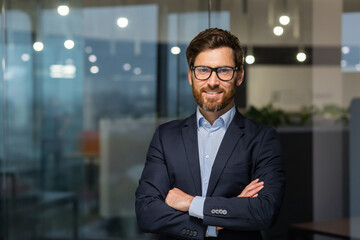 Portrait of successful mature boss, senior businessman in glasses and business suit looking at camera and smiling, man with crossed arms working inside modern office building. - obrazy, fototapety, plakaty