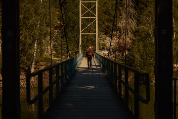 Fototapeta premium Man with backpack walking across a narrow suspension bridge over a river in a lush forest during golden hour. Cinematic view of outdoor adventure, hiking, and exploration in nature.