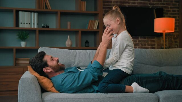 Happy father and funny little girl toddler teen kid resting on couch enjoying playtime laughing adult male parent playing with daughter tickle child spending free time together good family relations