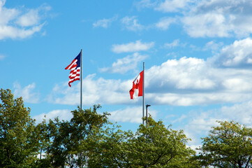 The flag of the United States of America and the flag of Canada flying in the sky above row of trees in northern new york on a summer afternoon.