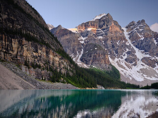 Sunset lights up the mountain peaks of the Three Sisters on the clear turquoise waters of Moraine...