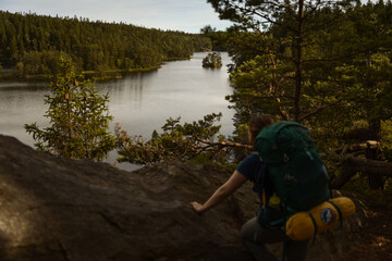 A woman hiker wearing a backpack standing at a lookout point in the forest watching over a lake in summer.
