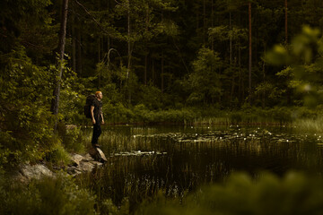 Fototapeta na wymiar A caucasian man wearing an orange beanie and a backpack standing on a rock by a lake in a forest.