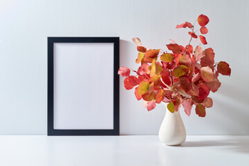 Mockup with a black frame and colorful autumn leaves in a vase on a light background. Empty poster frame mockup for presentation design, text, lettering