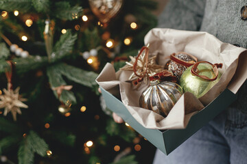 Woman in cozy sweater holding box with christmas baubles in atmospheric festive room. Decorating...