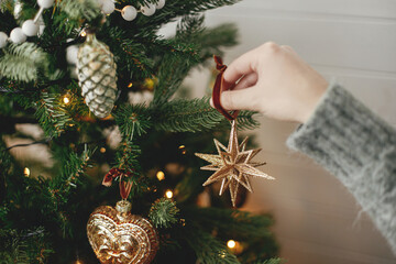 Hands decorating christmas tree with stylish bauble in atmospheric festive room. Merry Christmas! Winter holidays preparation. Woman in cozy sweater putting vintage star on tree