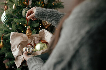 Stylish woman in cozy sweater decorating christmas tree with stylish bauble in atmospheric festive room. Merry Christmas! Winter holidays preparation. Decorating xmas tree with vintage toy