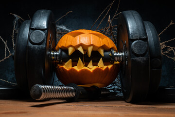 Scary carved Jack-o'-lantern Halloween pumpkin clenching teeth on a heavy barbell dumbbell. Gym...