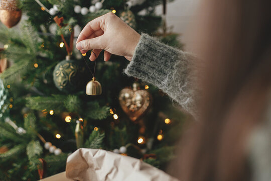 Hands decorating christmas tree with stylish bauble in atmospheric festive room. Merry Christmas! Winter holidays preparation. Woman in cozy sweater putting vintage bell on tree