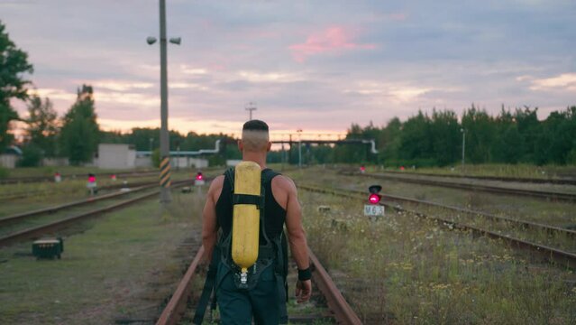 A muscular man with a cylinder drager compressed air on his back goes between the rails and semaphores glowing red. Back view