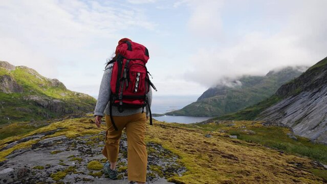 Girl with a big red backpack hikes through the highlands of Norway. The trail is barely marked. Fresh green grass. Taller mountains in the back covered partially with snow. Back view