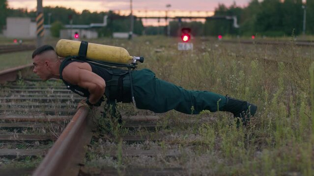 Young muscular man with liquefied air tank drager on his back is training outdoors and doing push-ups from the rail
