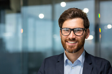 Close up photo portrait of successful and happy businessman, mature boss with beard and glasses working inside modern office building, senior investor smiling and looking at camera. - obrazy, fototapety, plakaty