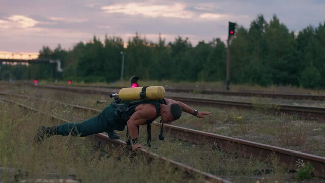 Young muscular man with drager balloon on his back is training on the railroad tracks and doing push-ups on one arm from the rail