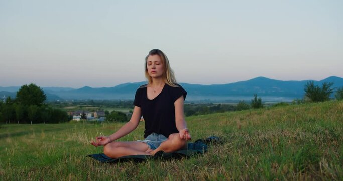 Woman practises yoga at sunrise on a meadow. Young spiritual girl meditates in lotus pose nature outdoors, slow motion. Concept of fitness, healthy lifestyle