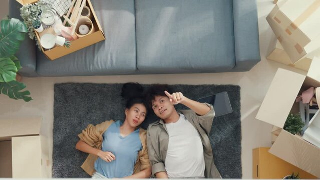 Top view. Happy Asian couple lay down and relax on carpet share idea for decoration furniture in empty area on living room at new house. Husband wife move house, Apartment Rent And Ownership Concept.