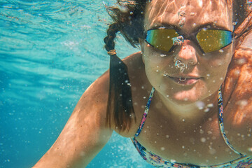 Young woman in reflective swimming glasses swim under water in sea, sun lit bubbles over her face, closeup underwater detail