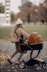 Happy family outdoors, a young stylish fashion mother holding baby near stroller in the autumn on nature.