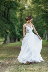Fototapeta premium Portrait of young attractive woman holding edges of white wedding dress, spinning outside in park in summer, dancing.