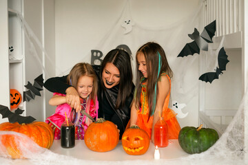 Cheerful friendly family is preparing for the celebration of Halloween. Mom helps her daughter carve a pumpkin. Children in the mother in festive carnival costumes. Table is decorated with cobwebs
