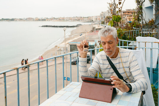 Senior grey-haired man using tablet device on terrace by the sea