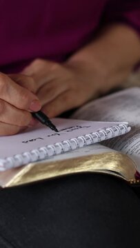 Female hand writing in notebook with a black marker with open Holy Bible Book in her lap. Vertical video. A close-up. Christian Scripture study biblical concept.