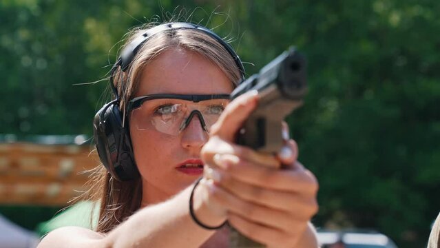 Front closeup outdoor portrait of focused powerful caucasian pretty girl in protective glasses and headphones aiming at target with black gun. Blurred trees in the background. High quality 4k footage