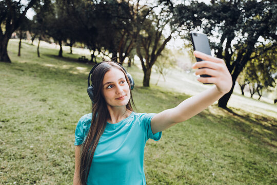 Smiling teenage girl wearing a turquoise t shirt and headphones, taking a selfie with her mobile in a natural space