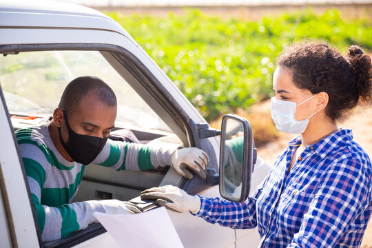 Hispanic female farmer in medical mask discussing contract with driver of transport company near car on farm field. Pandemic prevention and social distancing concept