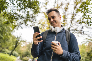 One young adult caucasian man walking in the city town near park using mobile phone smartphone texting sms or video call with cup of coffee happy male tourist standing alone real people copy space