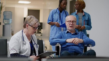 Old specialist talking to senior patient with disability, attending medical appointment with female caretaker and physician. Retired man wheelchair user at checkup visit. Handheld shot.
