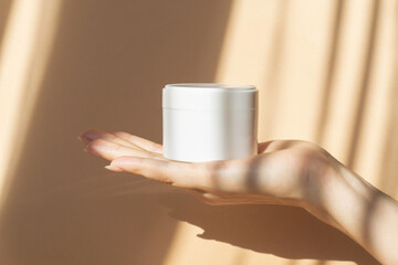 Mockup of white jar for packaging cosmetics on woman's hand. Unmarked container with moisturizer close-up on beige background, in rays of sunlight. Concept of skin care, rejuvenation