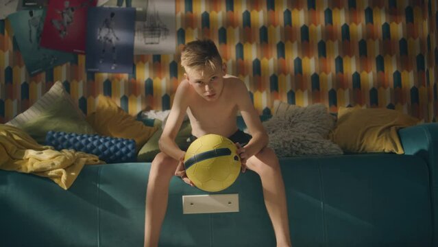 A teenage boy sits on a couch with a football in his hands. Shirtless young athlete boy. Focused for an upcoming soccer match. School sports team.