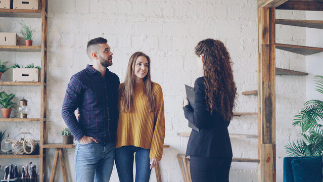 Attractive Young Couple Is Discussing Real Estate Deal With Female Realtor Standing Inside Beautiful House, Looking Around And Talking. Accommodation And People Concept.