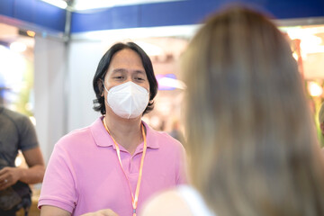 A male visitor wearing a face mask at an expo receives a brochure from a sales representative at...