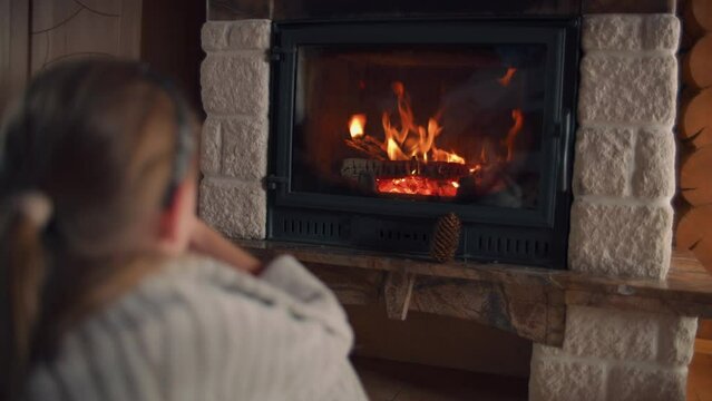 Young girl sitting by the fireplace on mild carpet, watching on fire and warming by coverlet in cold days, relaxing and spending leisure time at home. Back view.