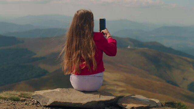 Young girl filming landscape of beautiful hills valley between mountains on phone, sitting on stone and making creative content at nature, spending leisure time outdoor. Back view.