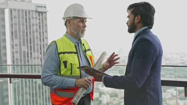 White caucasian professional construction architect holding an architecture blueprint plan discussing the project agreement details with his client an asian businessman on his digital tablet