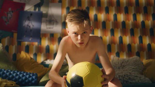 Thoughtful young boy sits on a couch with a yellow football in his hands. Shirtless young athlete boy. Focused for an upcoming soccer match. School sports team concept. Young athlete.