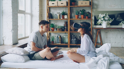 Married couple is having fun with augmented reality glasses, young woman is wearing then and smiling moving hands and her husband is using tablet. Modern technology and people concept.