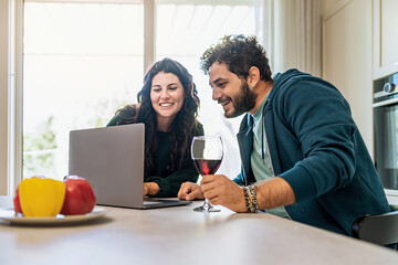 Happy couple of young newlywed using laptop in the kitchen surfing internet content and drinking red wine